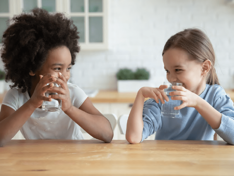 Two children smiling at each other while sitting at a table with glasses of water in their hands.