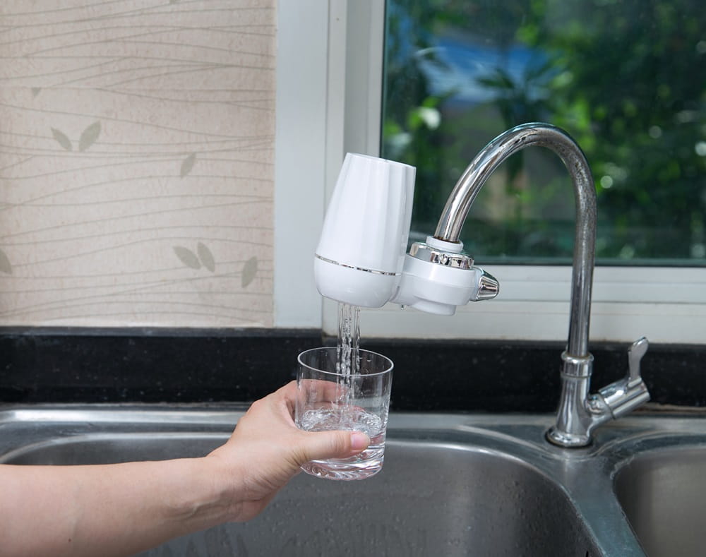 Hand holding up a glass of water to be filled from a kitchen faucet with an attached water filter