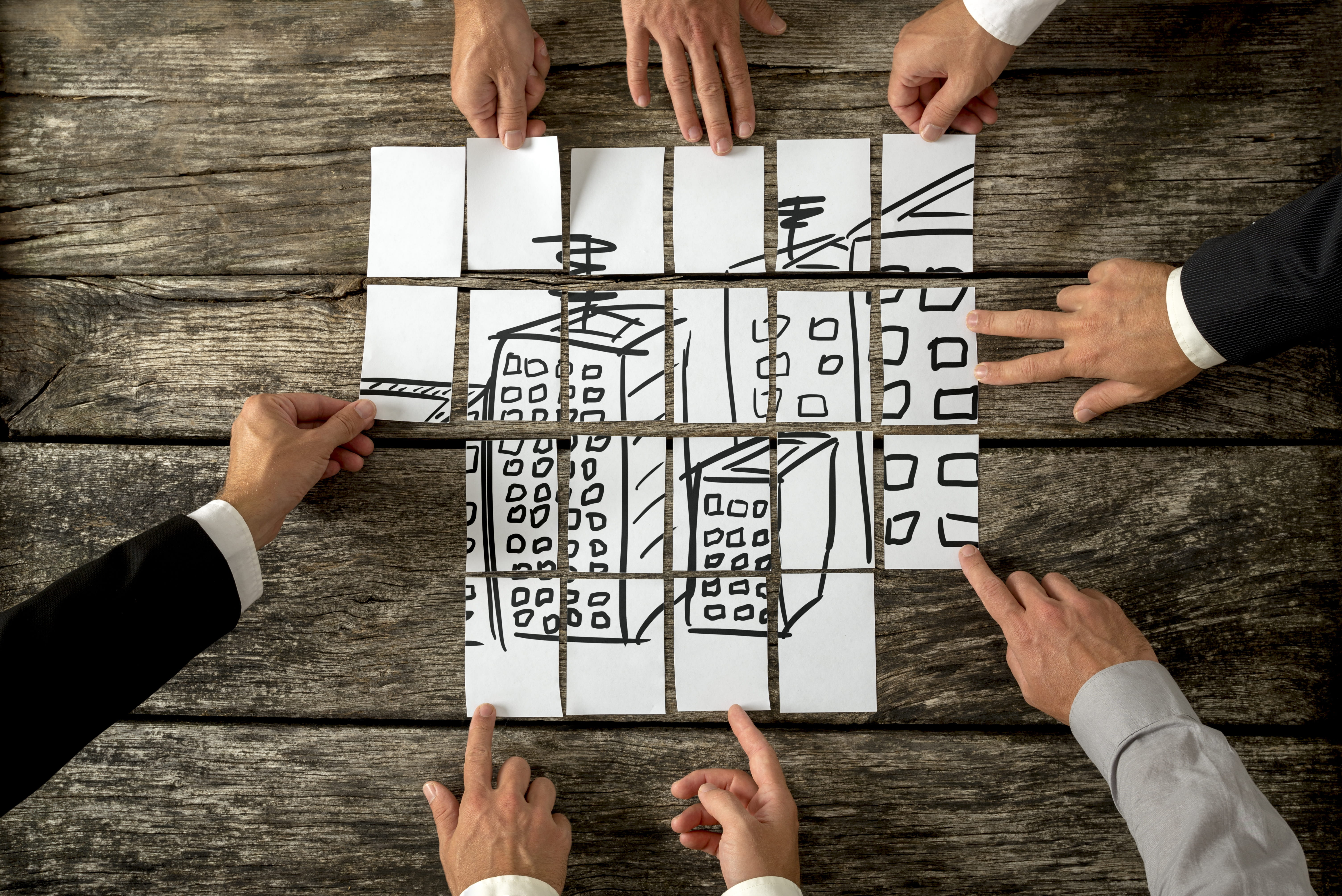 Hands putting pieces of paper together like a puzzle revealing a drawing of a set of buildings