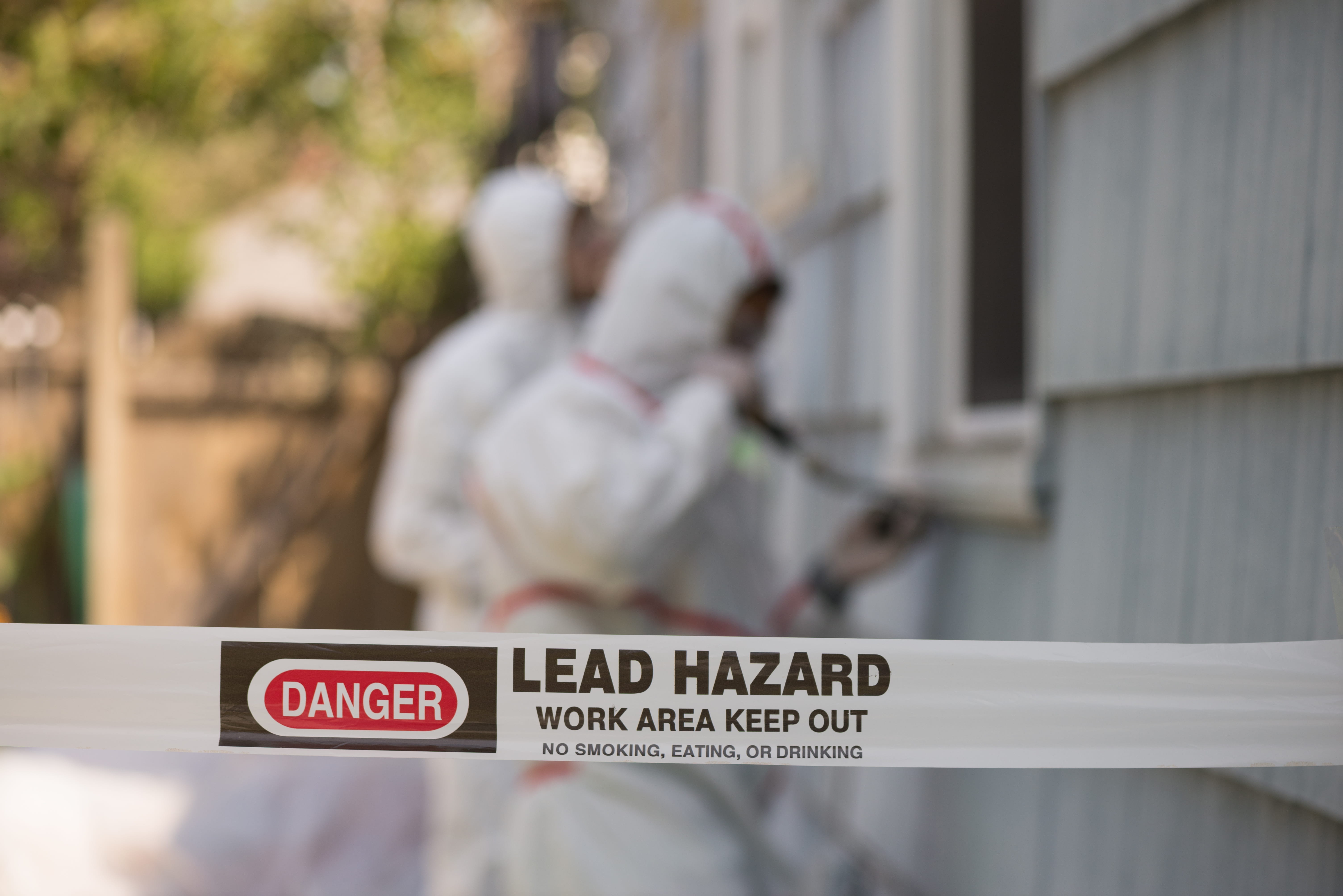Lead abatement workers removing lead based paint on the side of a house with caution tape in focus
