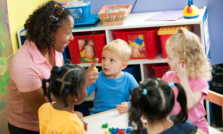 provider sitting at table talking to four children in classroom setting