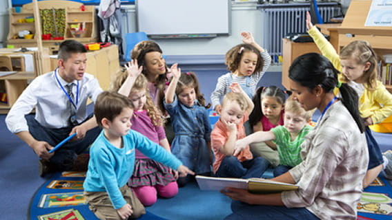 three teachers and eight preschoolers sitting on the floor while one teacher reads to them