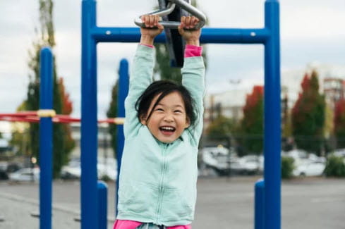girl in green jacket hanging from monkey bars