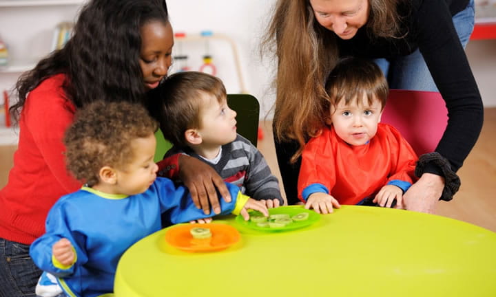 three toddlers sitting at a table eating in a care setting as two providers kneel over them helping