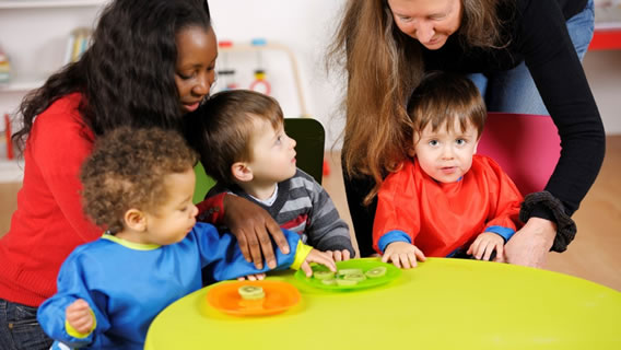 three toddlers sitting at a table eating in a care setting as two providers kneel over them helping
