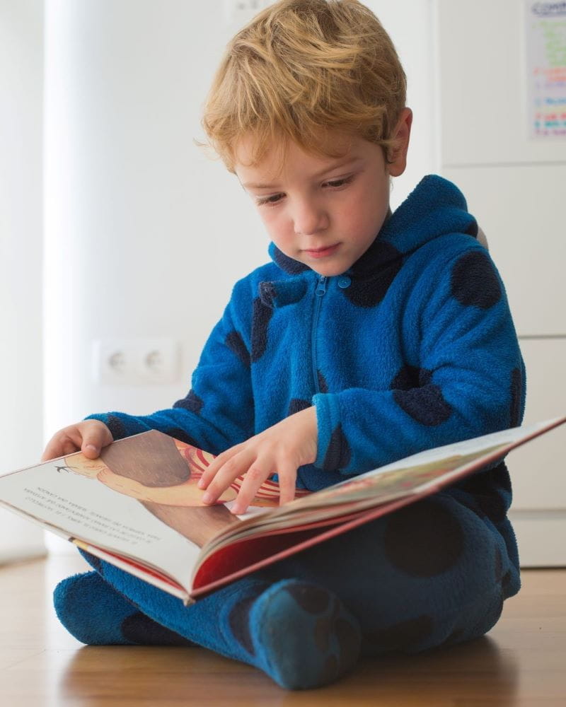 A young child with blonde hair and a blue hoodie sits on a wood floor reading a book.