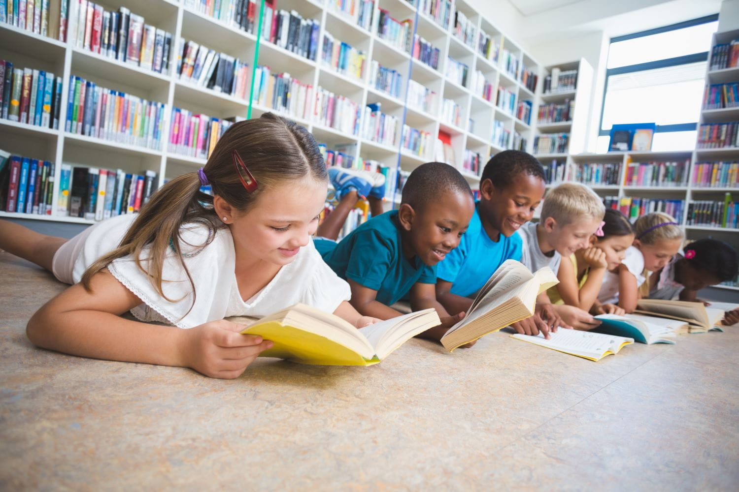 Children laying on the floor in a library reading books. A bookshelf is visible in the background.