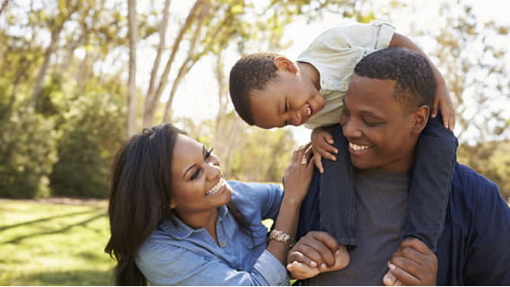 mother outside holding shoulder of father who is holding smiling preschooler on his shoulders