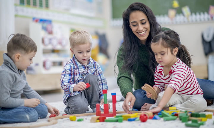 preschool teacher sitting on classroom floor with three preschooler playing with blocks