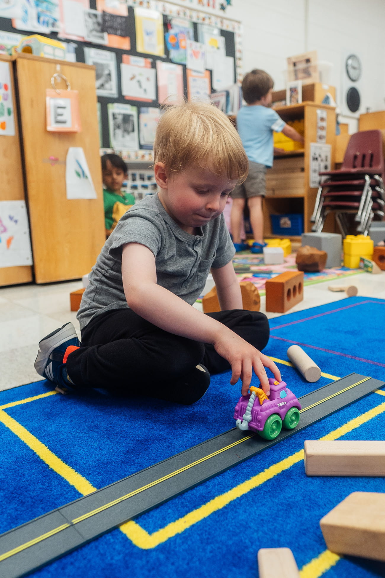 A close up of a child playing with a toy car on the carpet in a preschool classroom setting