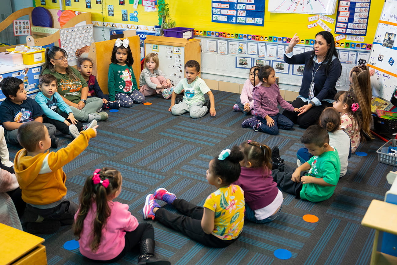 Preschool students sitting in a circle on a rug with their teacher in a classroom setting