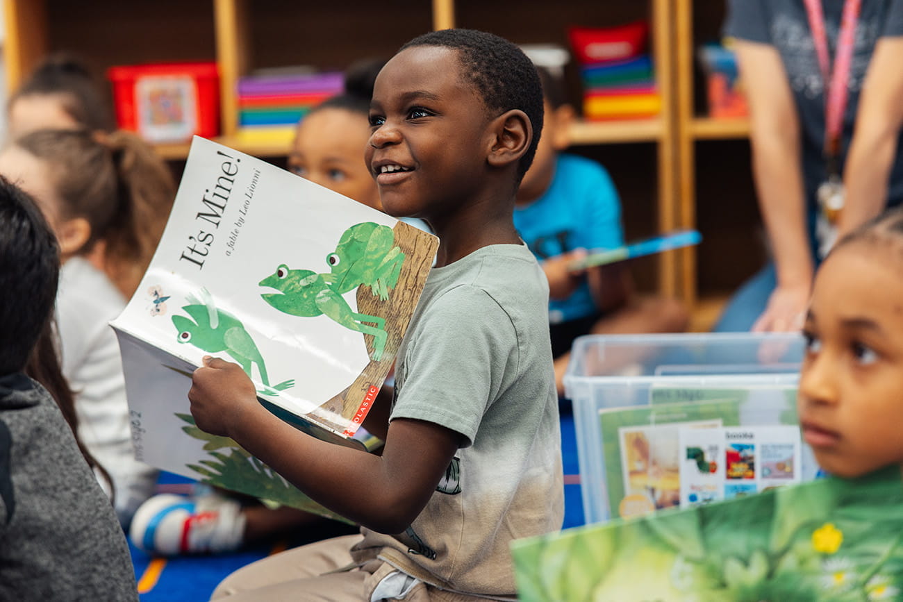 Child holding a book and smiling at the camera in a preschool classroom setting