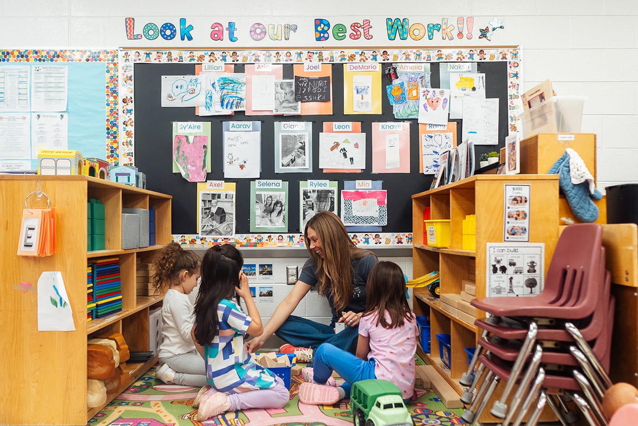 A teacher sitting on a floor surrounded by students in a preschool classroom
