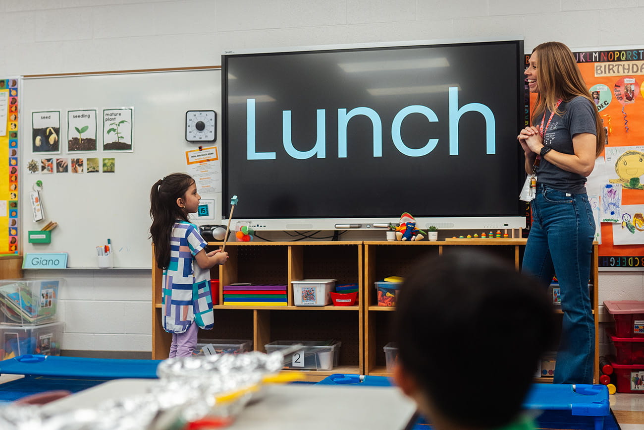 A student at the front of the classroom with the preschool teacher pointing at a screen with the word LUNCH