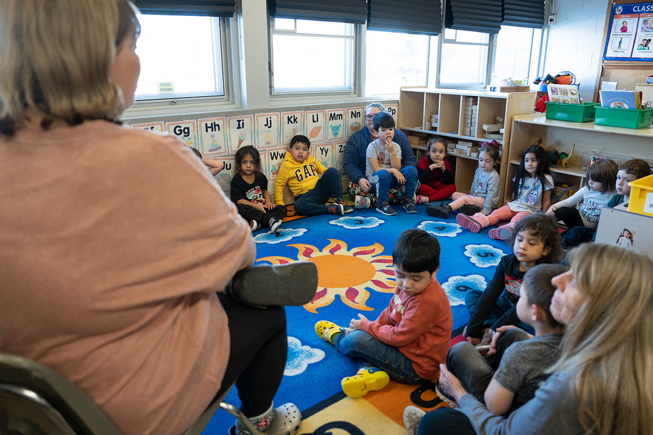 A teacher sitting in a chair in front of students seated on a rug in a preschool classroom