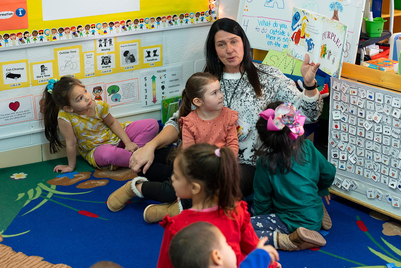 A teacher seated on a rug and surrounded by children in a preschool classroom