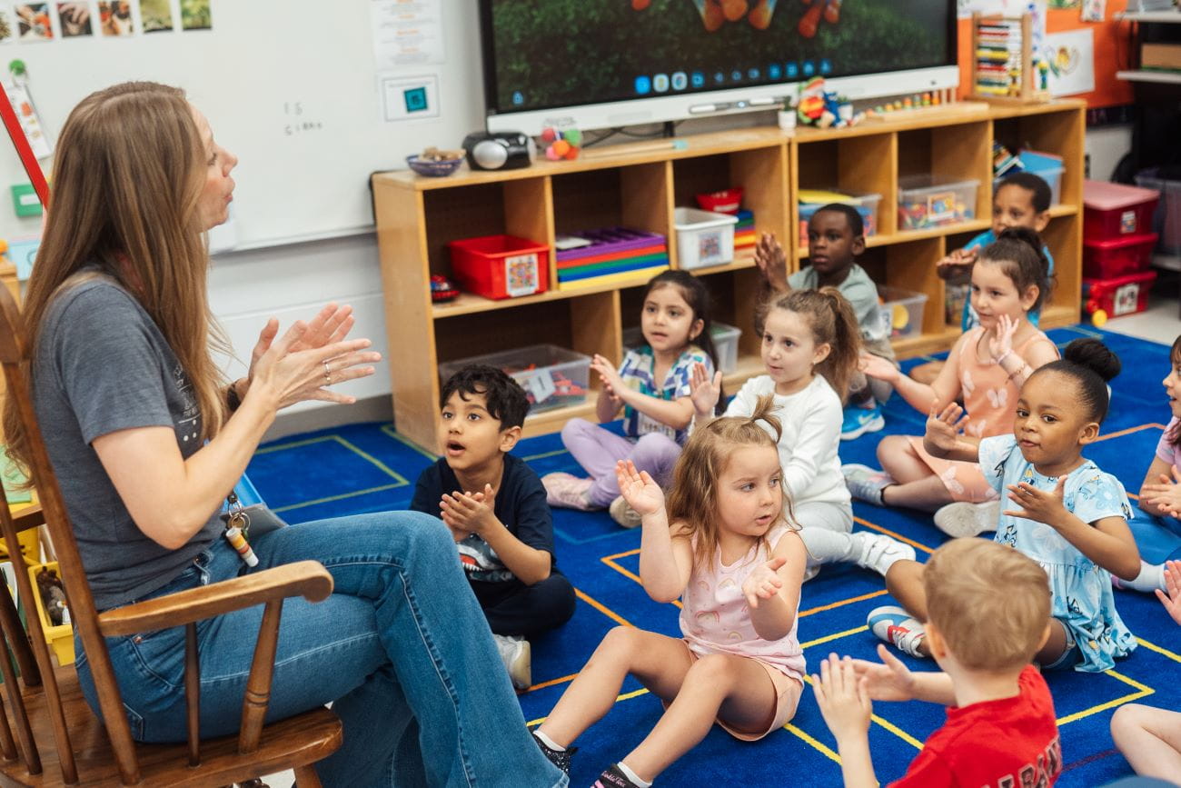 A teacher sitting in a rocking chair talking to students sitting on the carpet in a preschool classroom