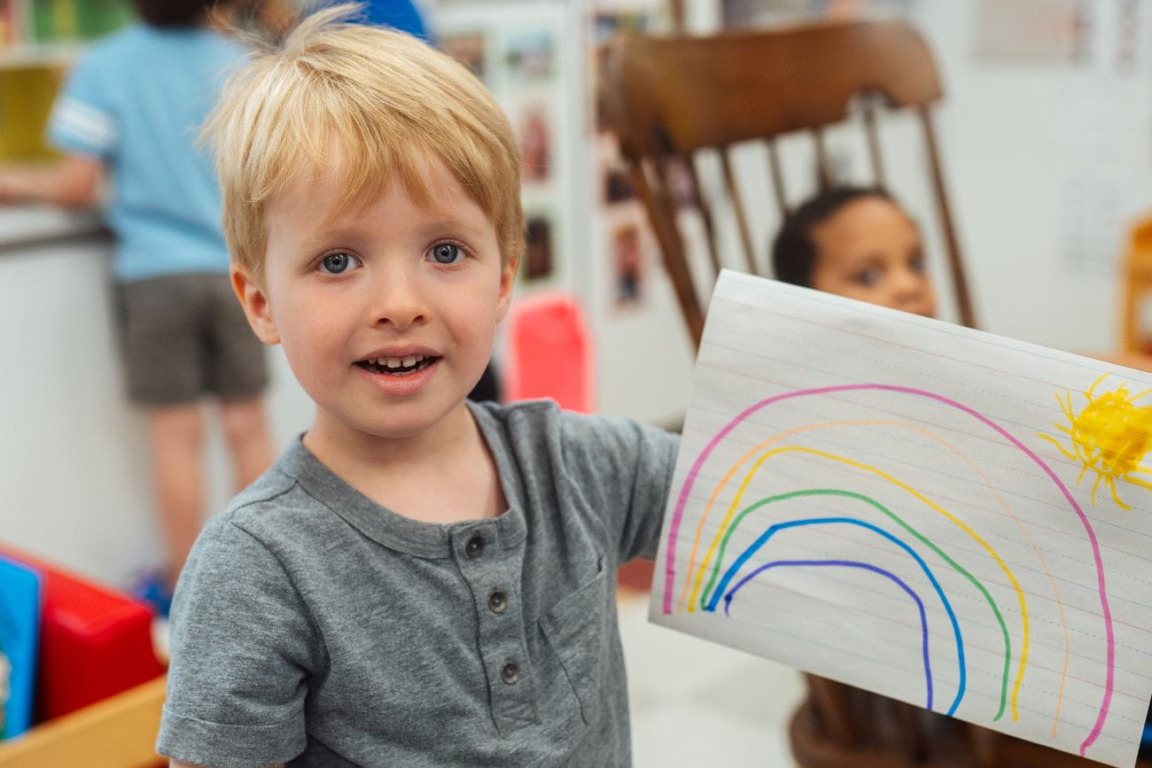 An up-close photo of a young child showing off his drawing of a rainbow
