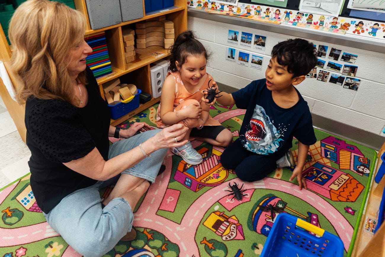 A teacher and two young students sitting on a rug in a preschool classroom