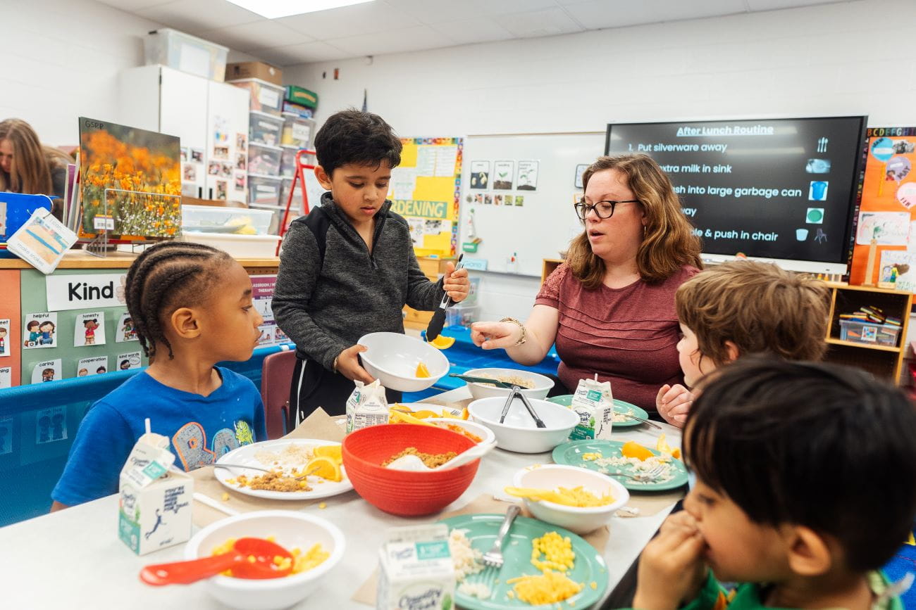 A teacher and young children sitting down to eat at a table in a preschool classroom