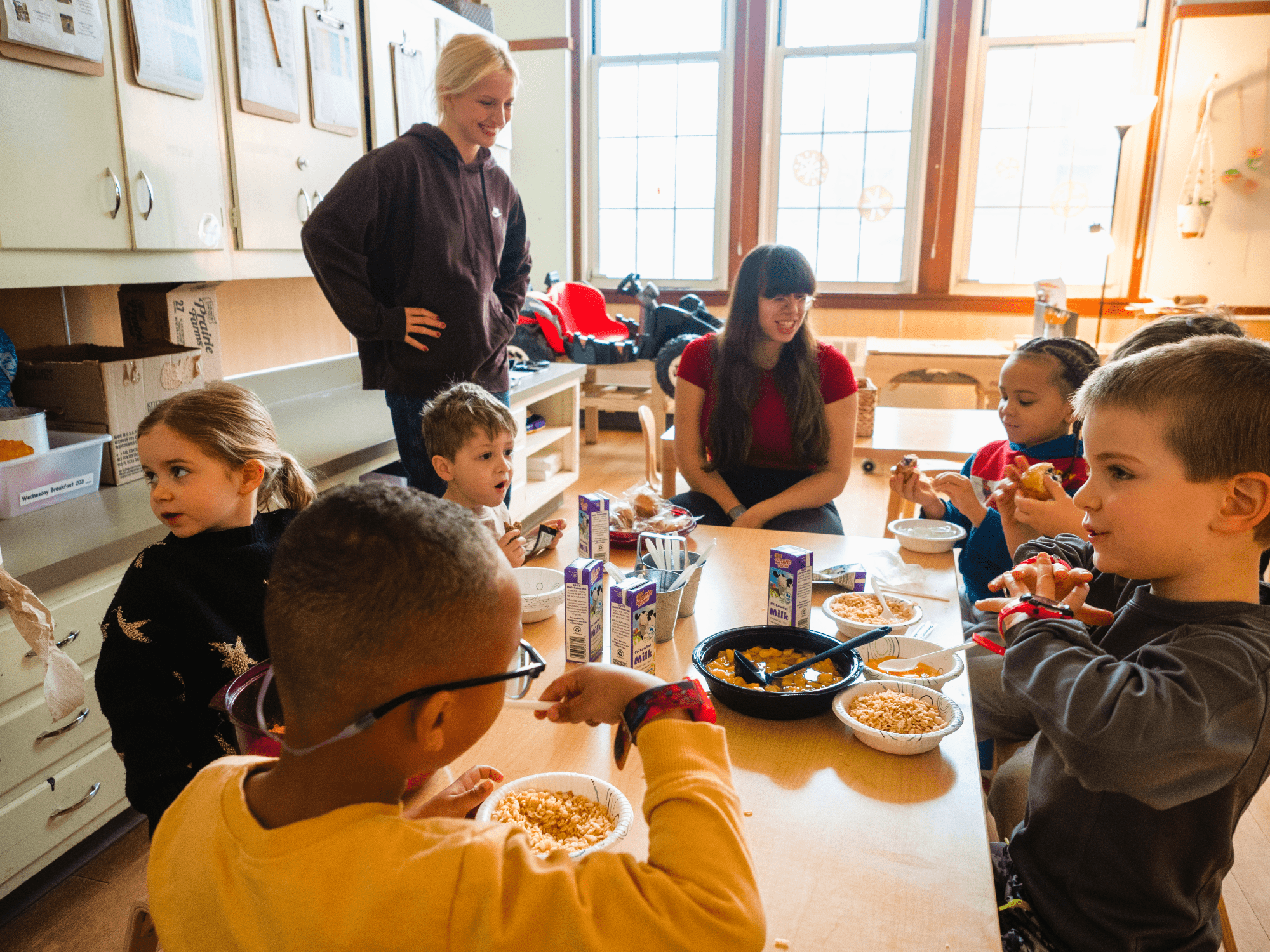 Children eating lunch at a preschool table with caregivers seated and standing nearby.