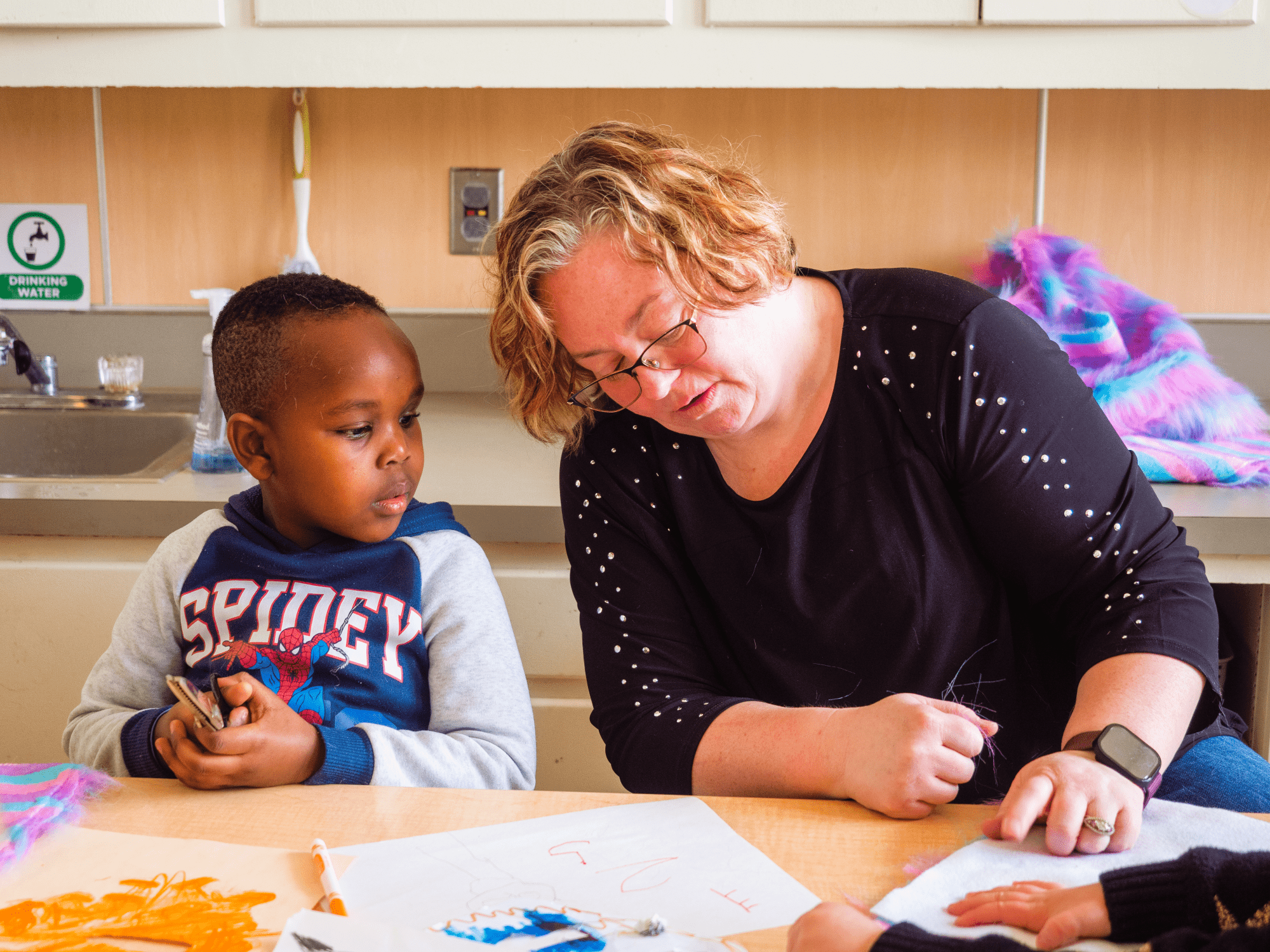 A teacher and a young student seated together at a table in a preschool classroom