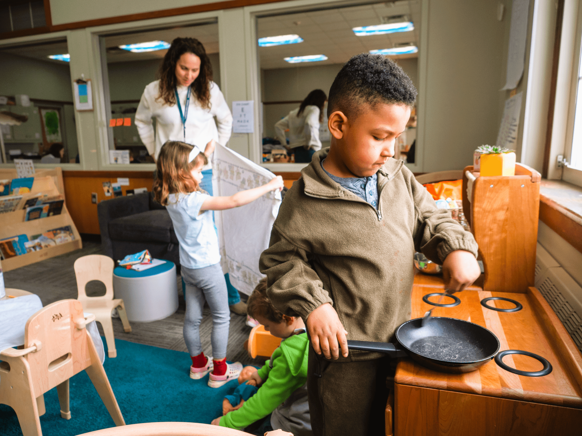 Preschool students playing in a toy kitchen area in a classroom while a caregiver watches