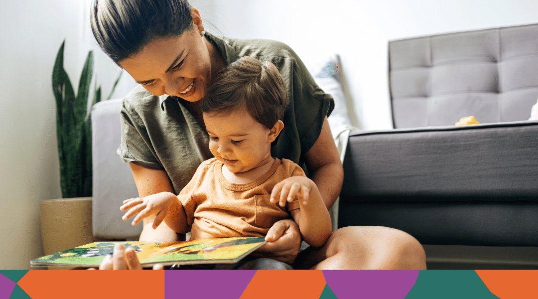 A young child sitting in an adult's lap on the floor looking at a book in a living room setting.