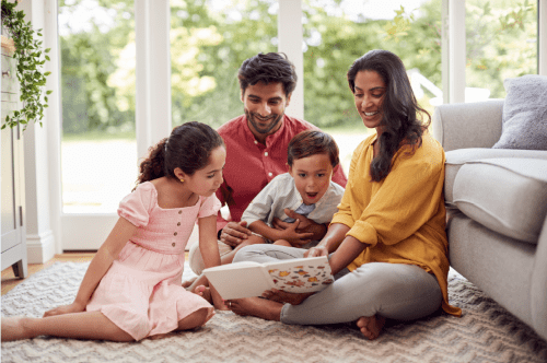 Two adults and two children sitting on the floor looking at a book in a brightly lit living room setting
