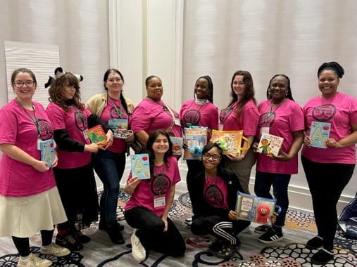 A group of women in pink shirts holding books pose together at the 2026 Literacy Day of Learning event.