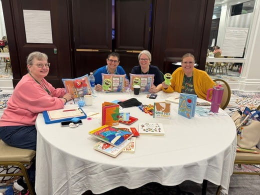 Four early educators display children's books and training materials they received during the Literacy Day of Learning session.