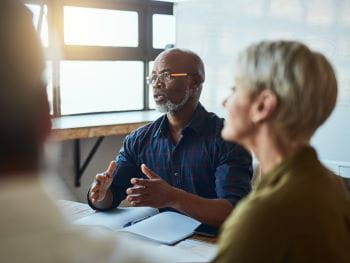 A man addresses people sitting at a conference table in an office setting