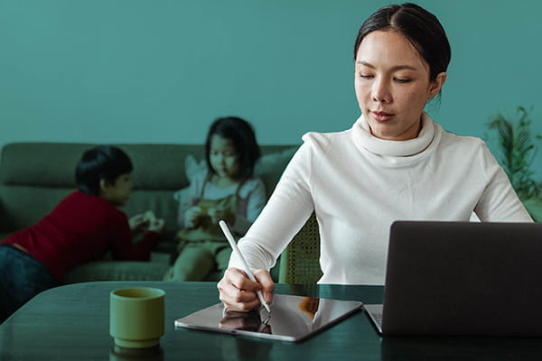 A women sitting at a table working on her laptop and tablet while kids are sitting on the couch in the background.