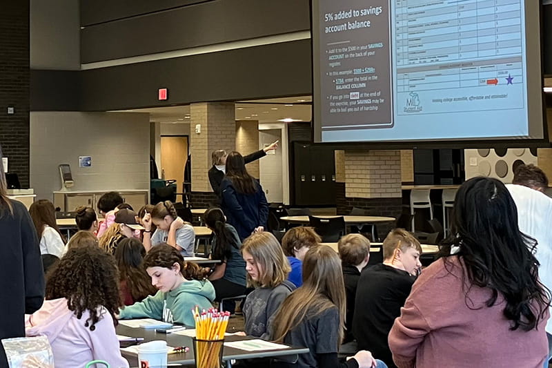 A Department of Treasury employee gives a financial literacy presentation on budgeting to a cafeteria filled with middle school students