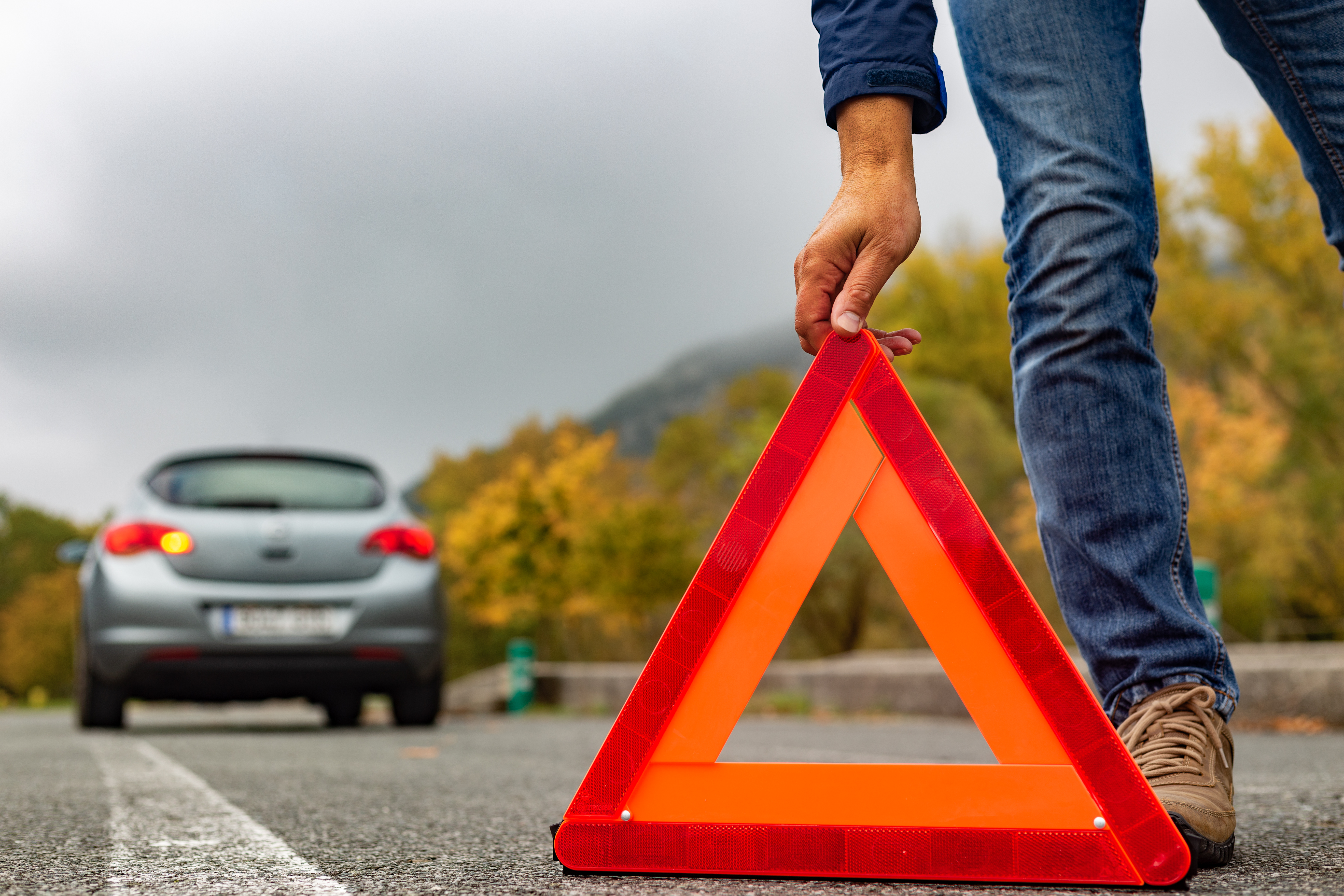 A person putting a hazard triangle down with their vehicle in the background.