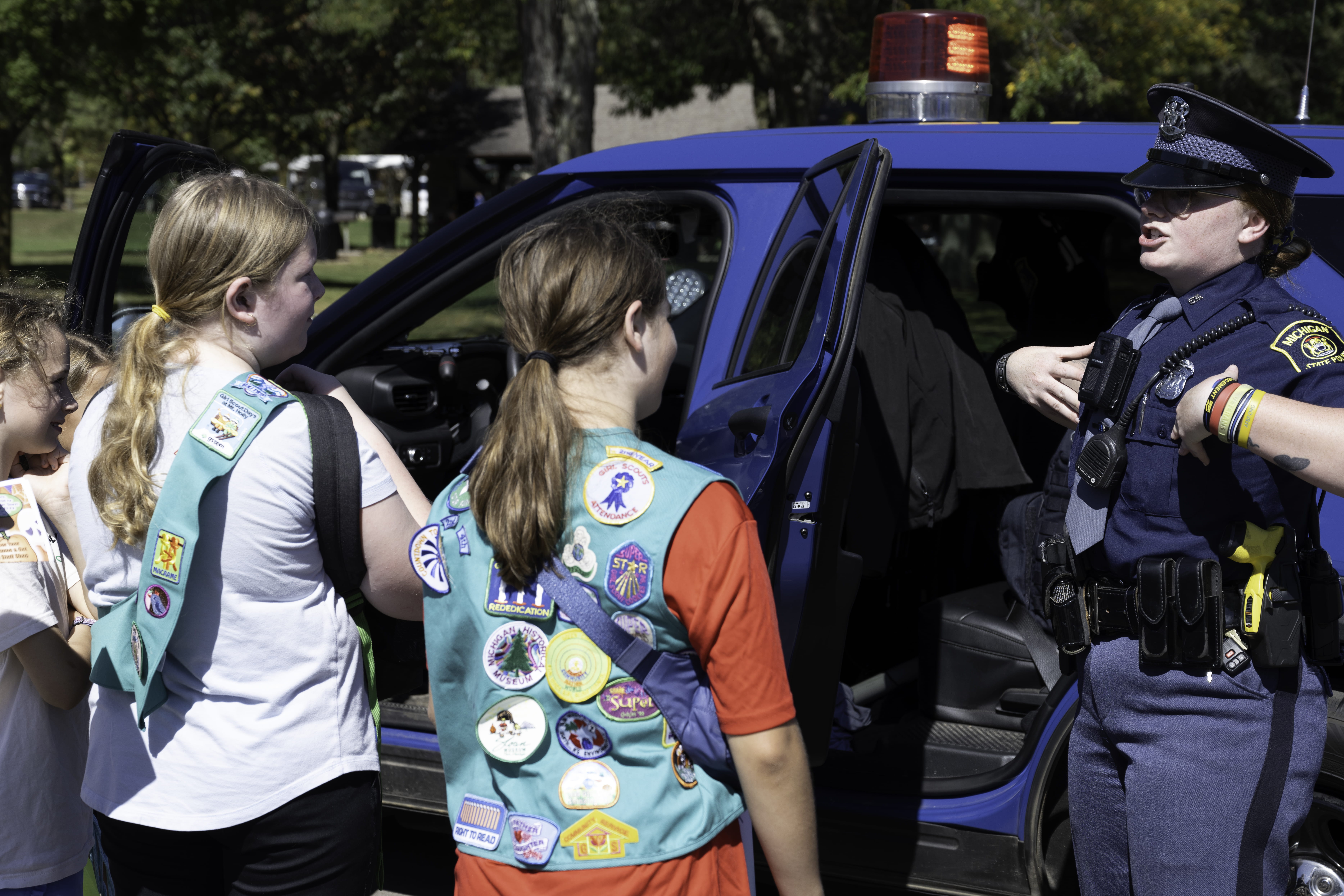 Two young girls wearing green girl scout vests  and patches learning from an MSP trooper while standing next to a patrol vehicle.