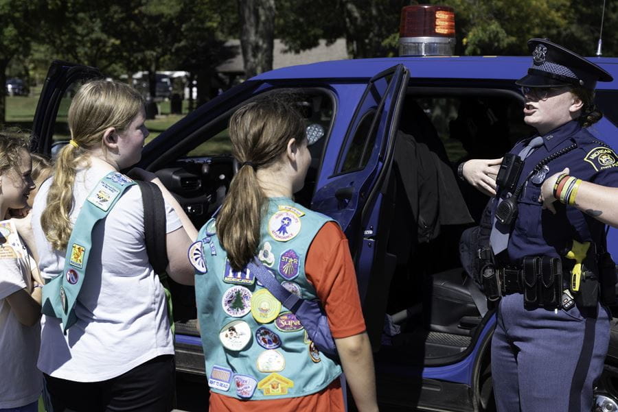 Two young girls wearing green girl scout vests and patches learning from an MSP trooper while standing next to a patrol vehicle.