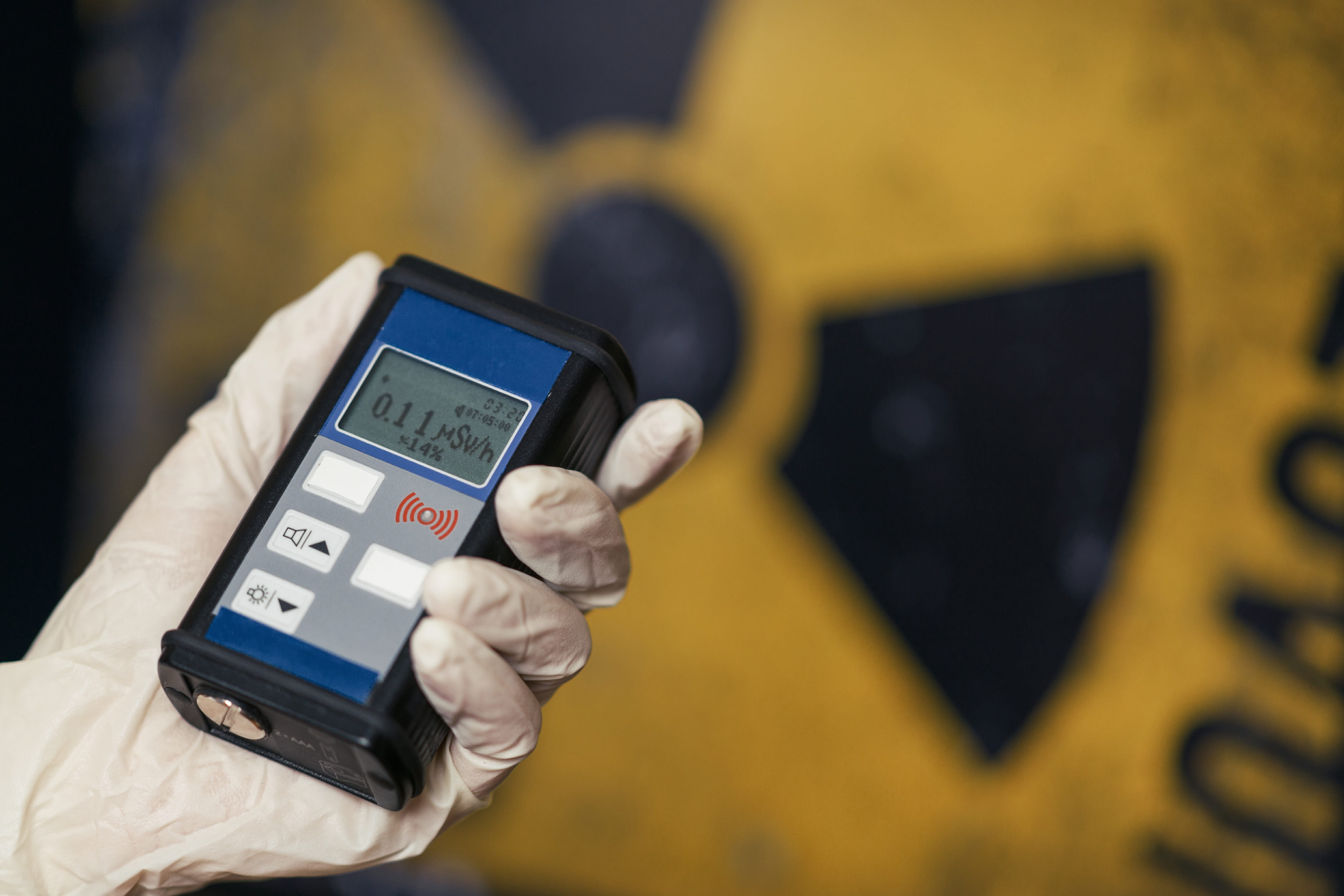 A hand holding a radiation detector with a radiation warning sign in the background.