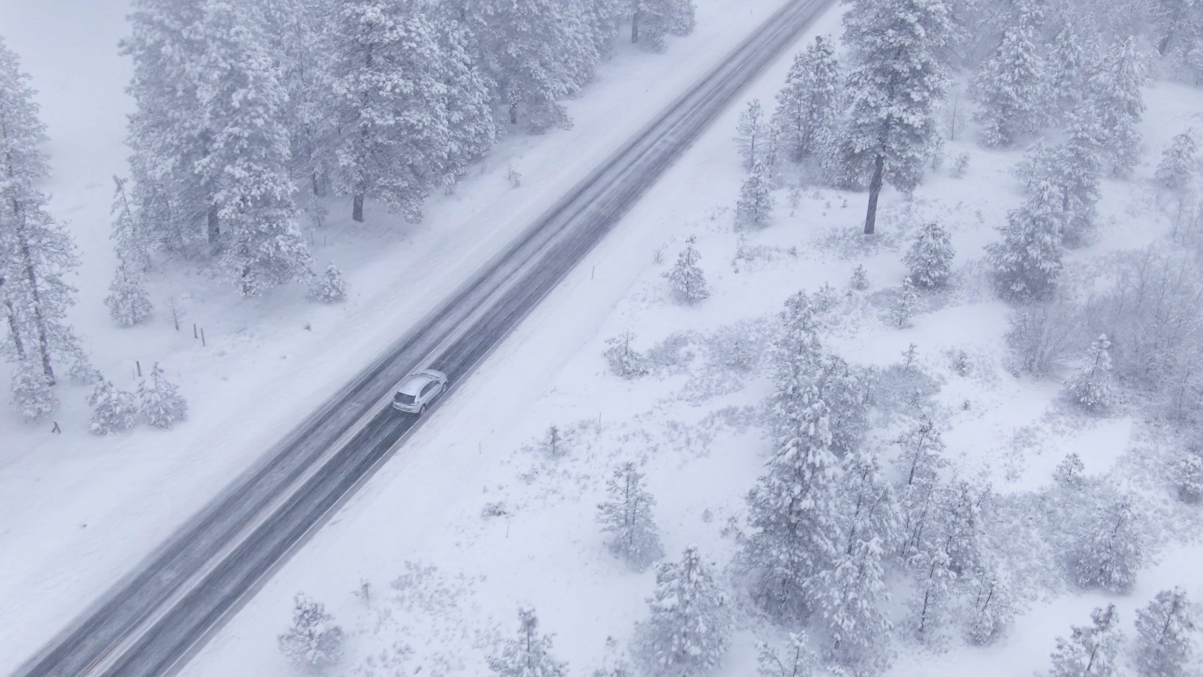 An aerial view of a car driving down a road in winter weather with snow and snow-covered trees on either side of the road.