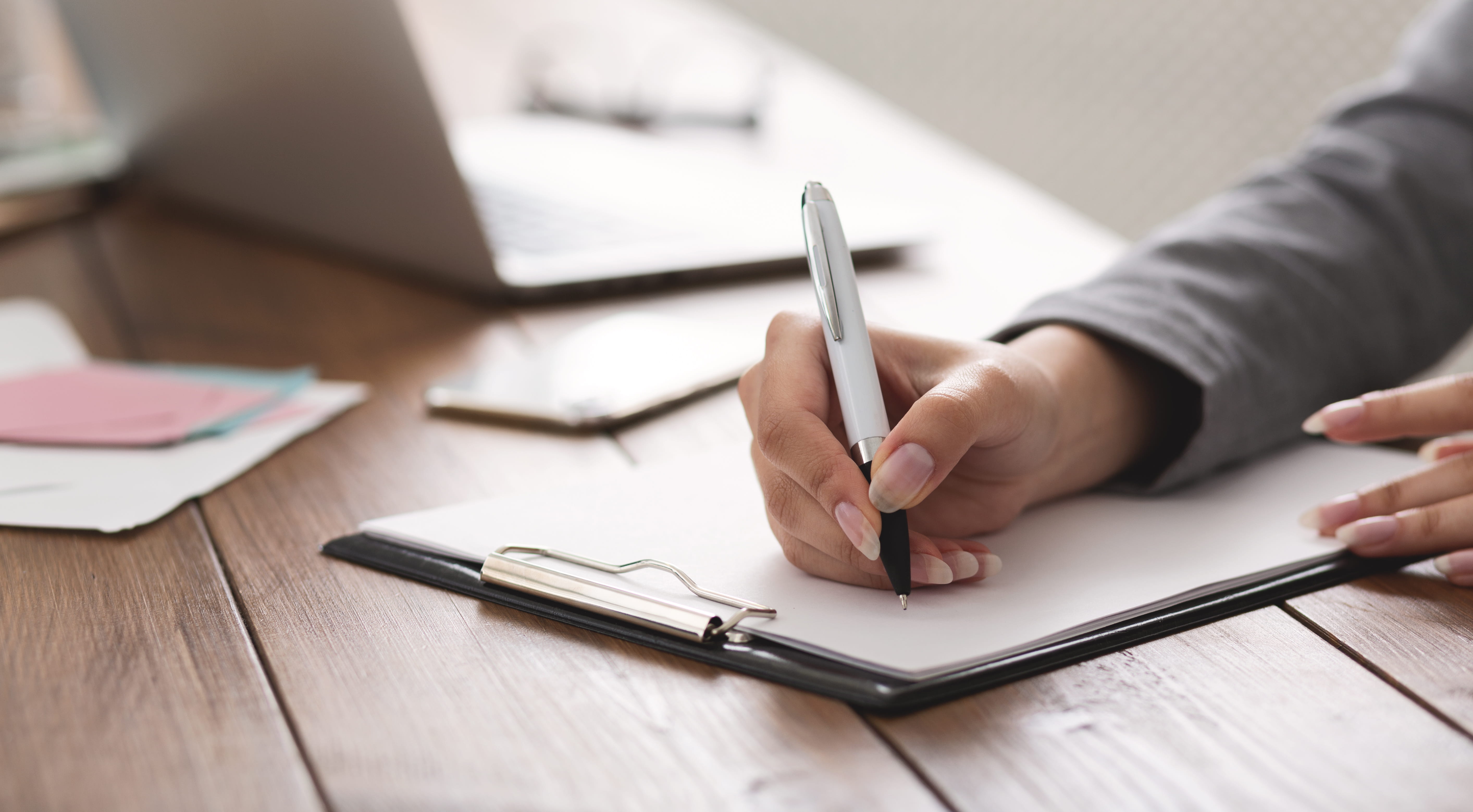 a person writing on a clipboard with a laptop in the background