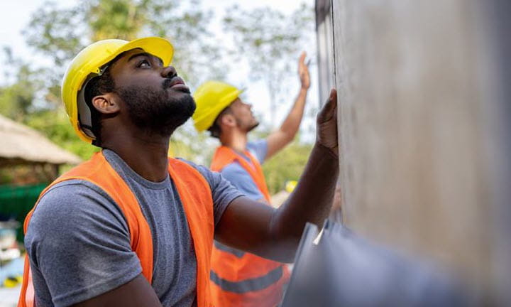Two construction workers wearing hard hats and vests work on the side of a wall structure.