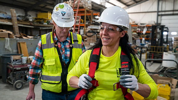 a woman in construction gear trying on a harness while another woman in construction gear instructs and watches her