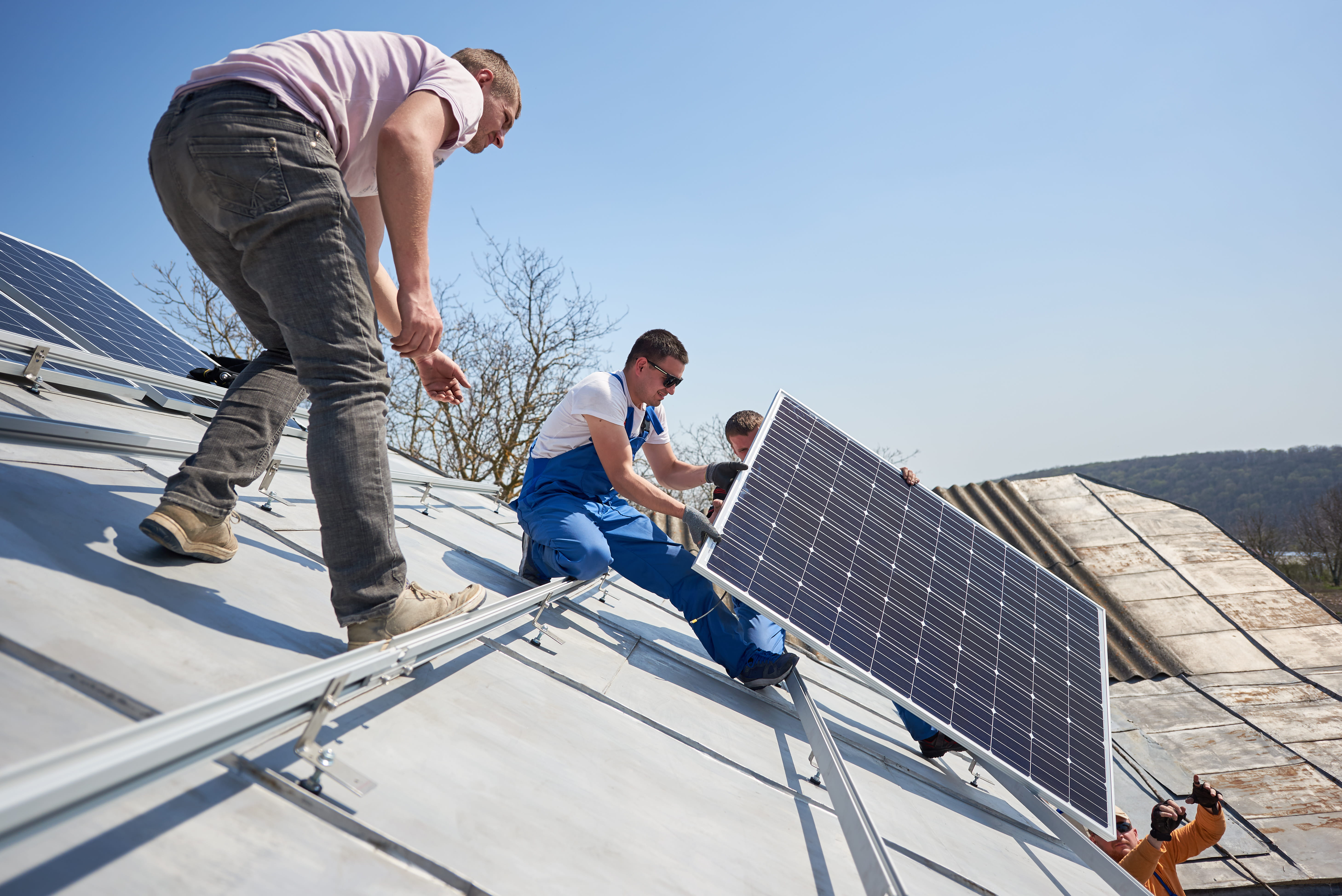 Electricians lifting blue solar module on roof of modern house. 