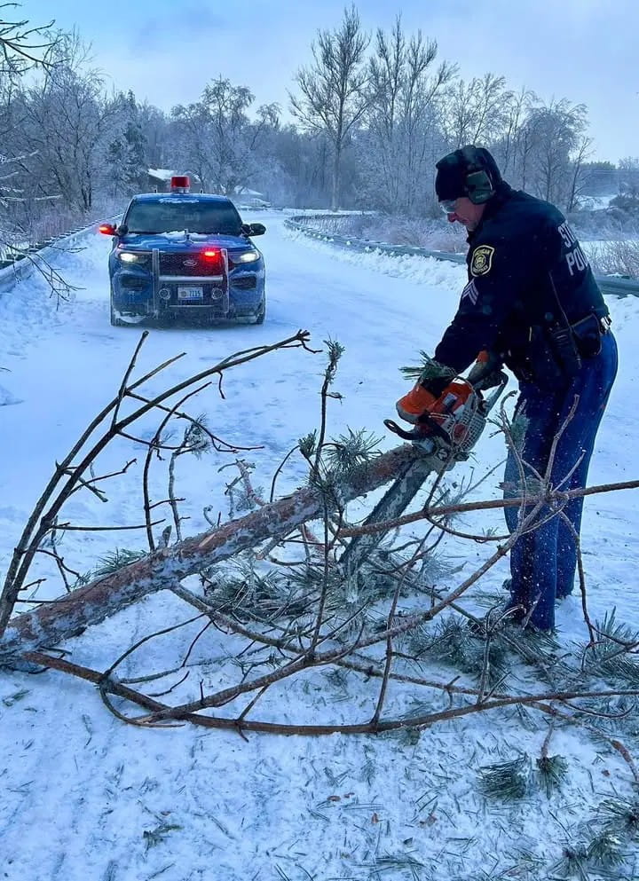 A Michigan State Police trooper using a chain saw to cut a large tree branch that has fallen across a snowy, icy road. 
