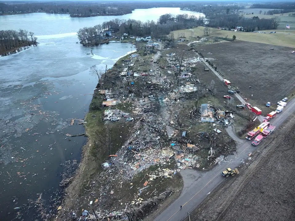 An aerial view of a lake with damaged buildings, trees, and debris from a tornado next to it. 