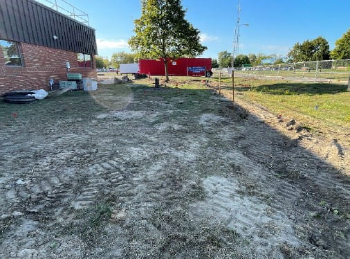 The lawn with dirt and construction vehicle tracks next to the current building. Construction materials are stacked next to the building.