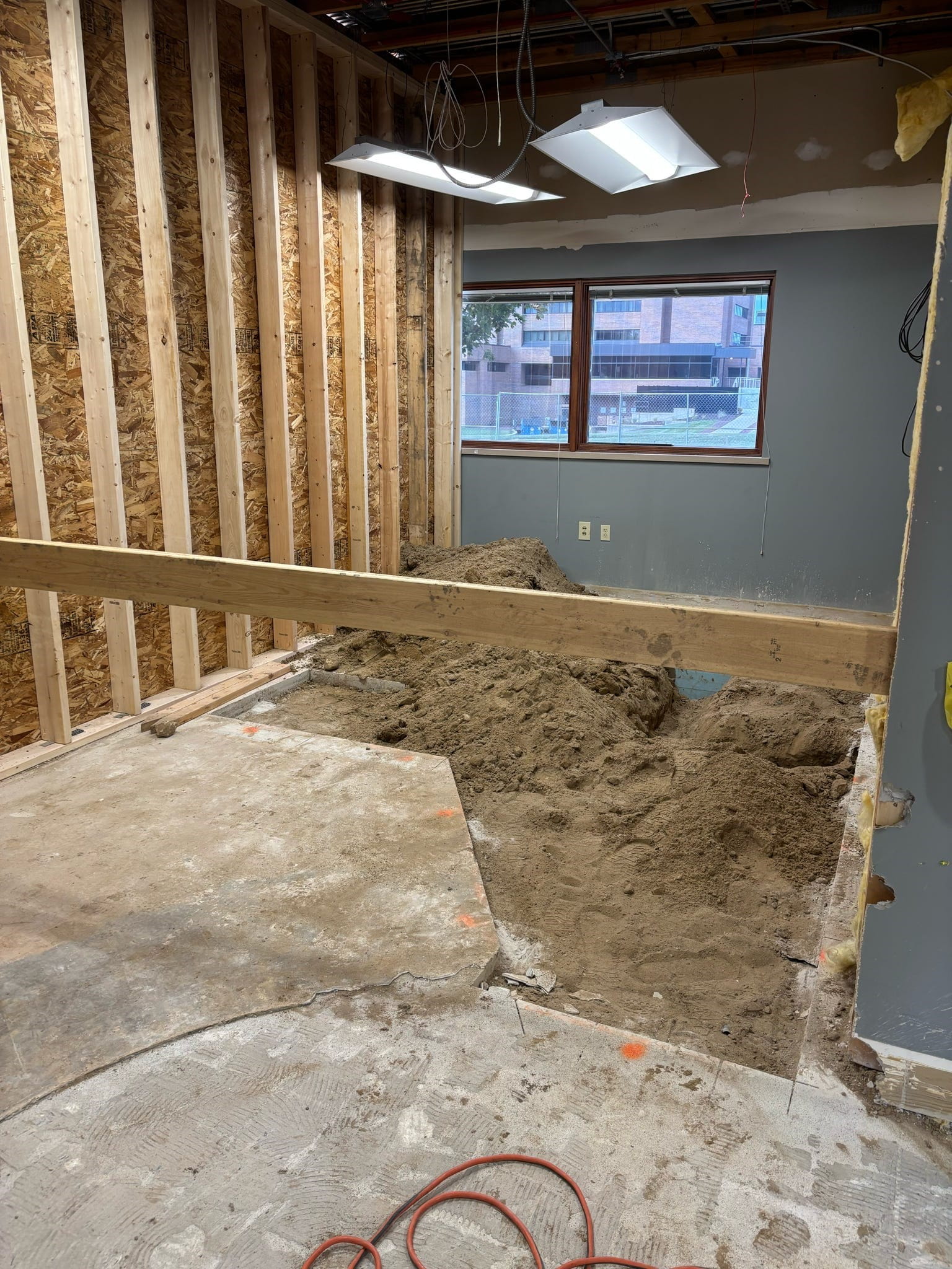 Inside the building, a room with exposed studs on one wall. Part of the concrete floor is removed and dirt is in it