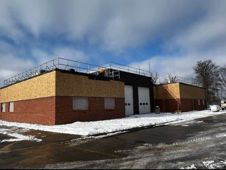 The outside of a building that is red brick on the bottom and plywood around the top. There are two white garage doors in the middle of the building. There is snow on the ground around the building. 