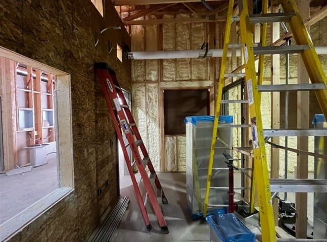 The inside of a room with exposed walls and a cutout for a window. There are ladders and building equipment in the middle of the room. 