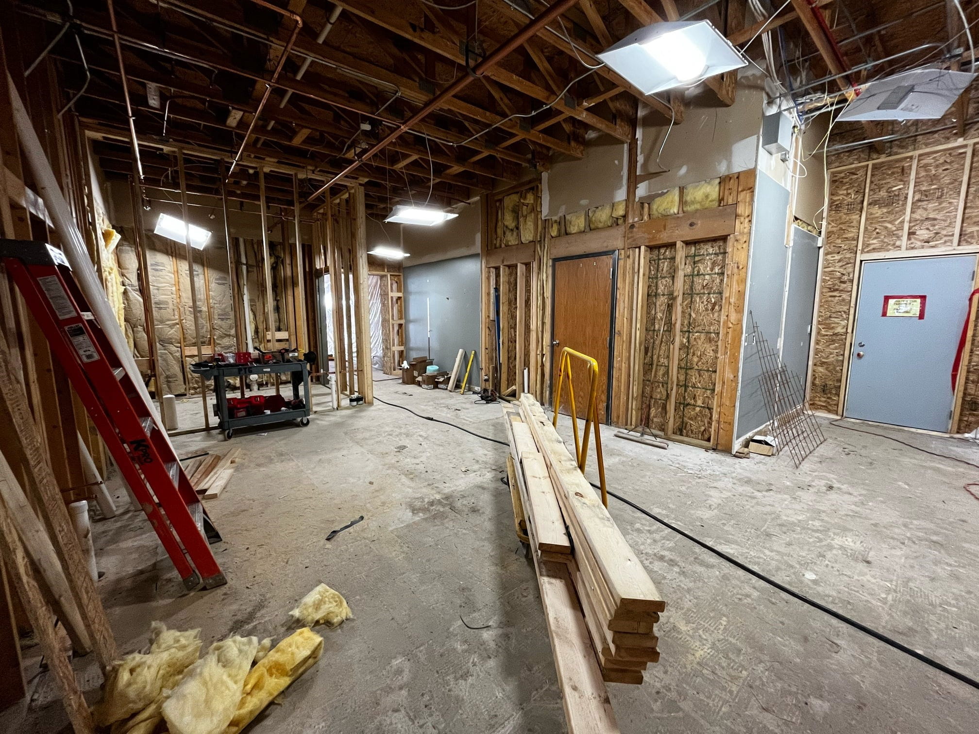 The inside of the existing building with exposed ceiling, walls, and concrete floor, with building materials and tools scattered about. 